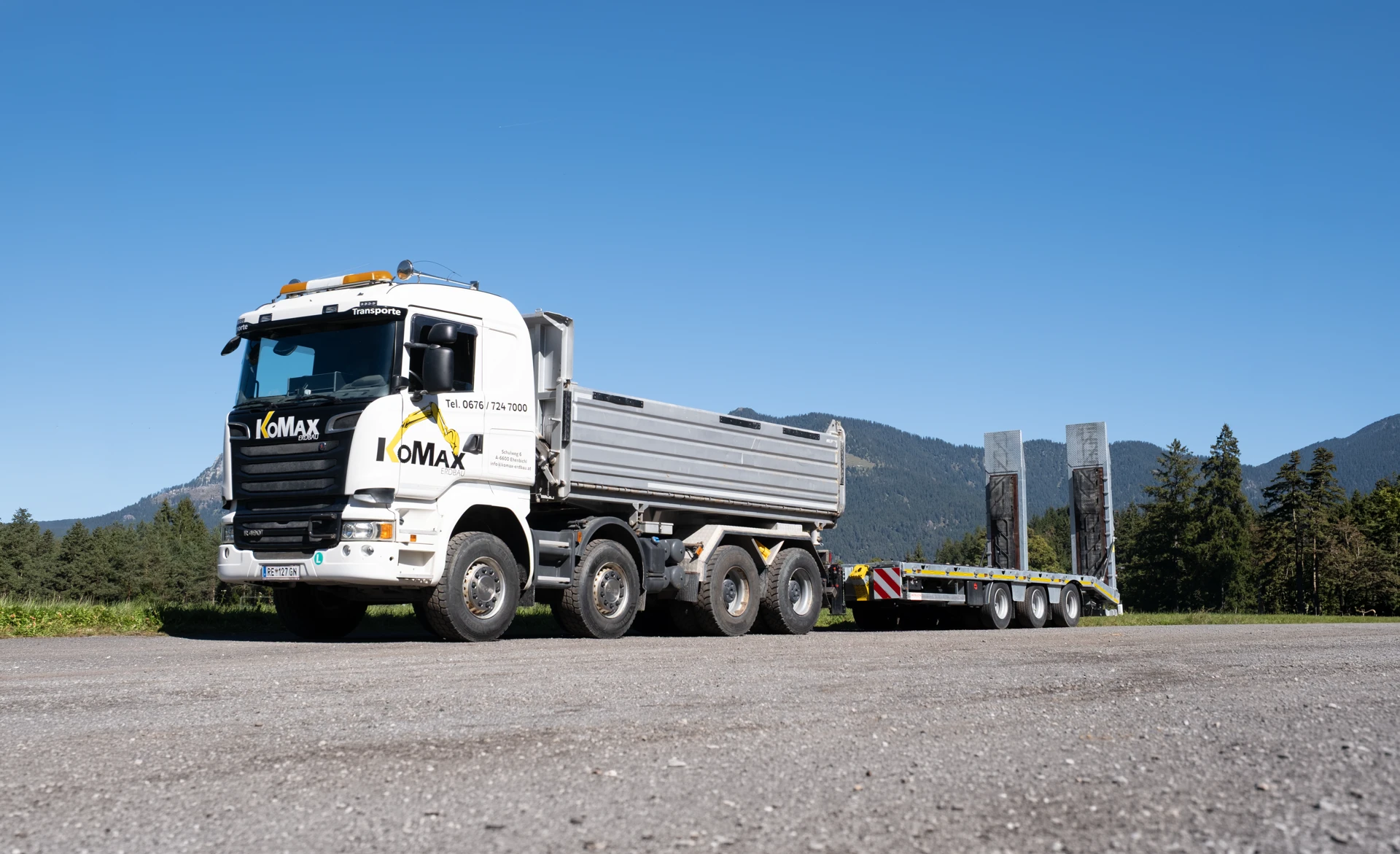 Weißer KoMax-Lkw mit Kipper und Tieflader-Anhänger auf einem Schotterplatz vor Wald und Bergen