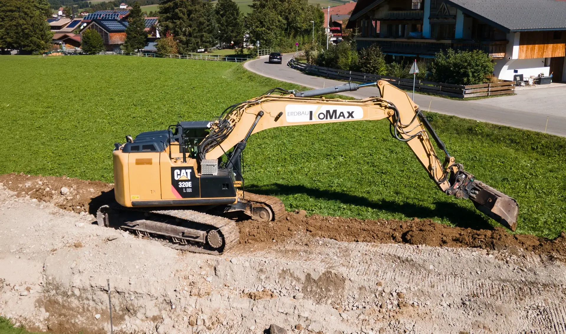 Gelber Cat-Kettenbagger mit KoMax-Schriftzug und Tieflöffel am Rand einer Baugrube, dahinter Wiese, Straße und Häuser