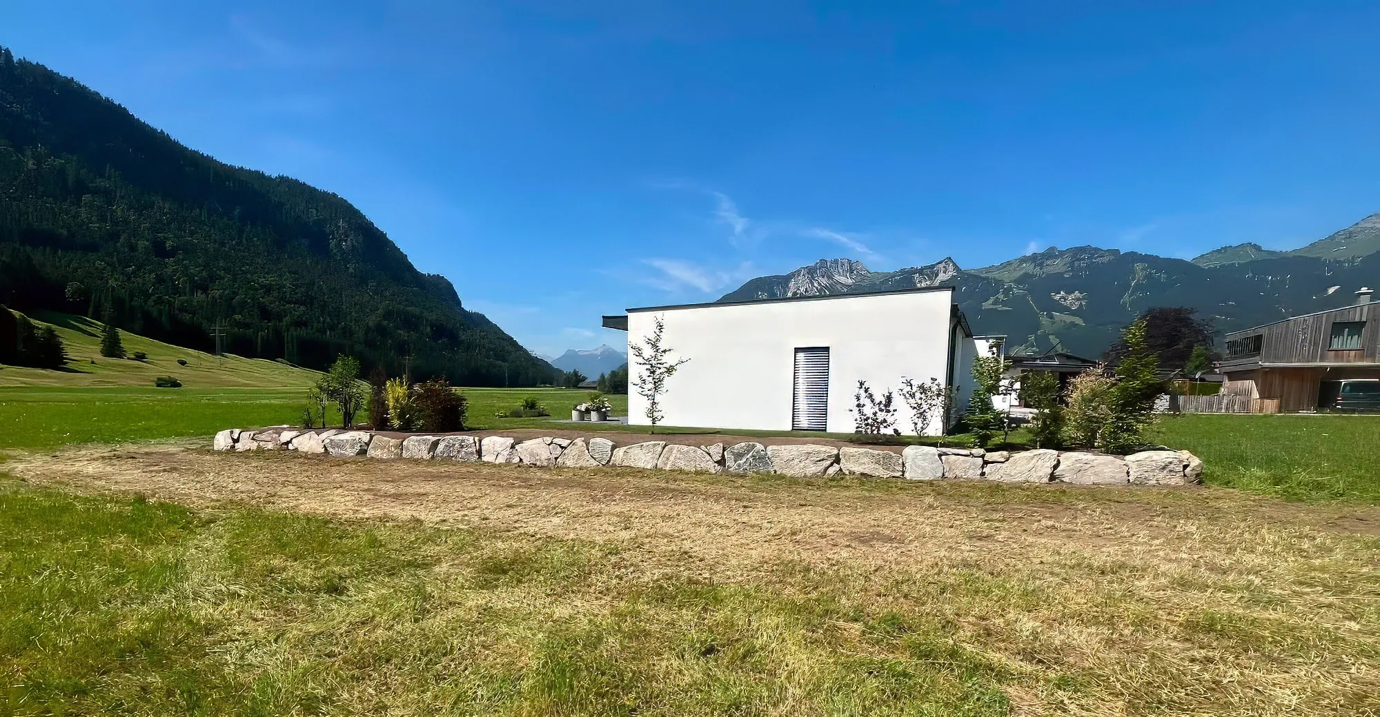 Neue Natursteinmauer aus großen hellen Steinen vor einem weißen Haus, dahinter Wald und Berge