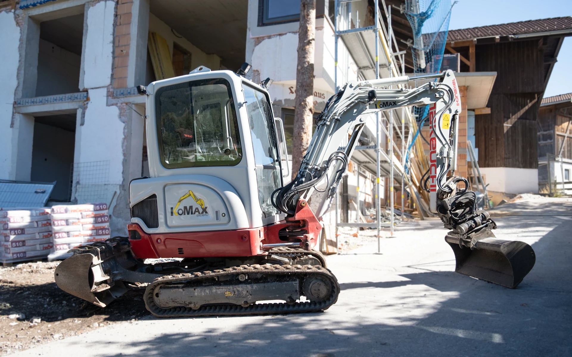 Weiß-roter Minibagger mit KoMax-Logo und Grabenräumlöffel vor einem eingerüsteten Rohbau