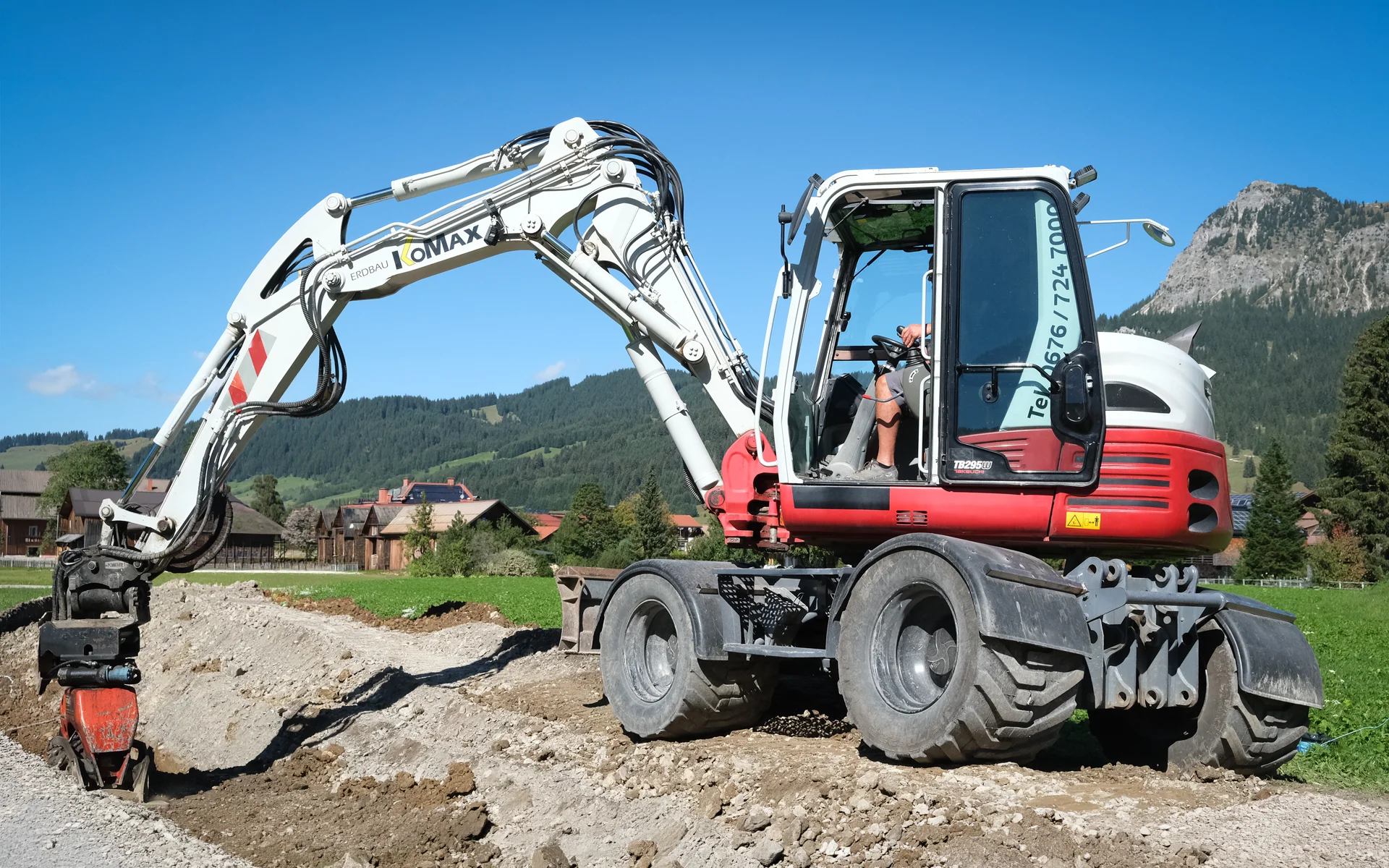 Weiß-roter Mobilbagger mit KoMax-Schriftzug am Ausleger und Anbauverdichter an einem frisch aufgeschütteten Graben, dahinter Wiese, Holzhäuser und Berge