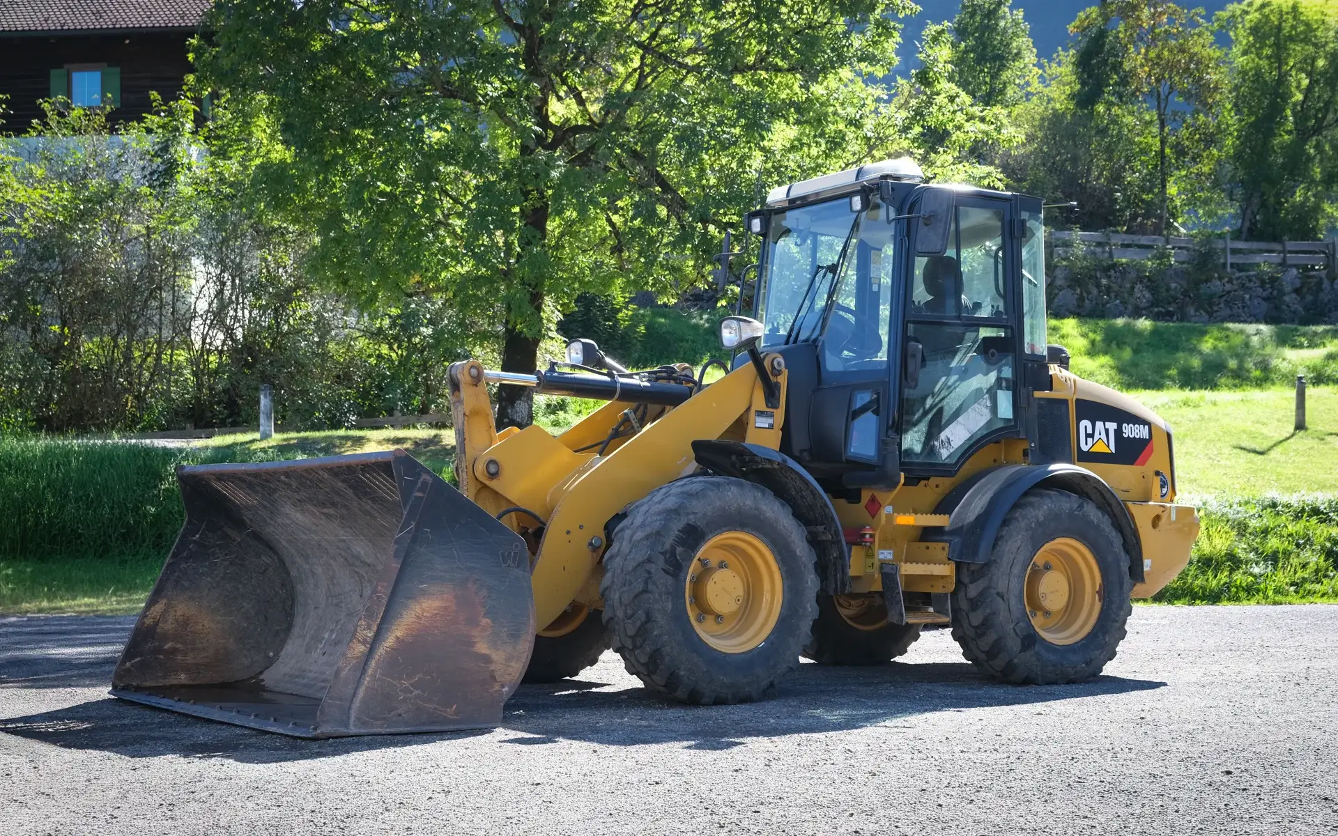 Gelber Cat-Radlader mit Schaufel auf einem Kiesplatz vor Bäumen und Wiese