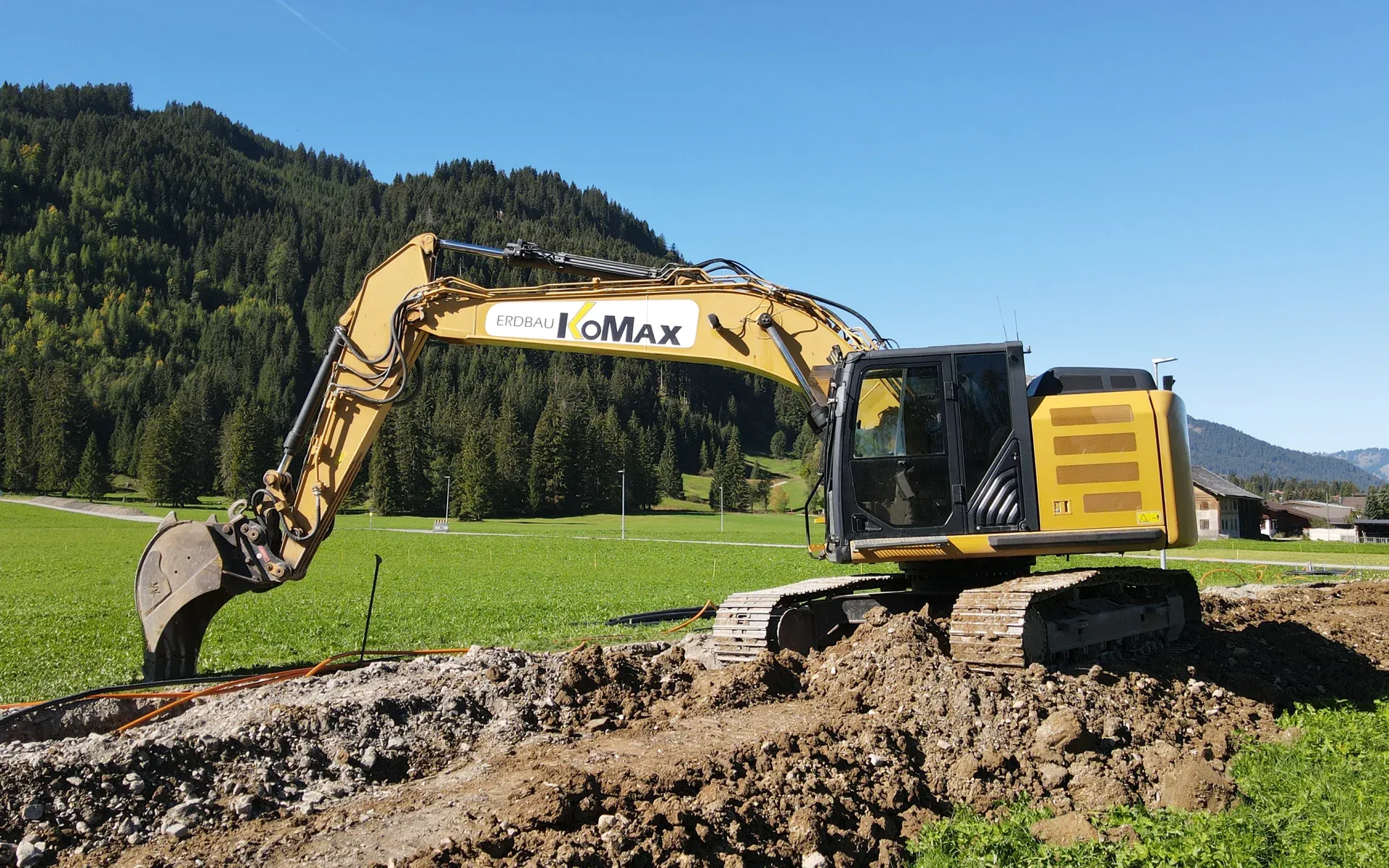 Gelber Kettenbagger mit KoMax-Schriftzug am Ausleger auf aufgeschüttetem Erdreich, dahinter Wiese und bewaldeter Berg