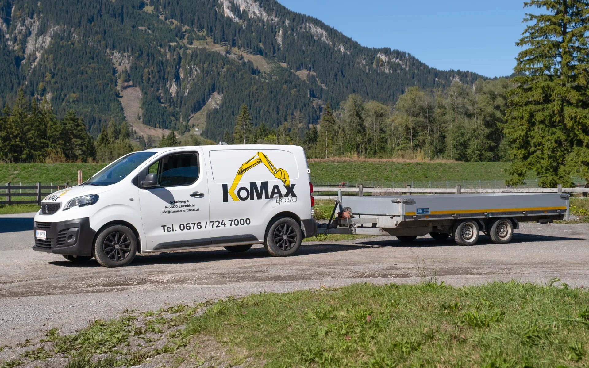 Weißer Transporter mit KoMax-Logo, Adresse und Telefonnummer, dahinter ein Pritschenanhänger, im Hintergrund bewaldete Berge