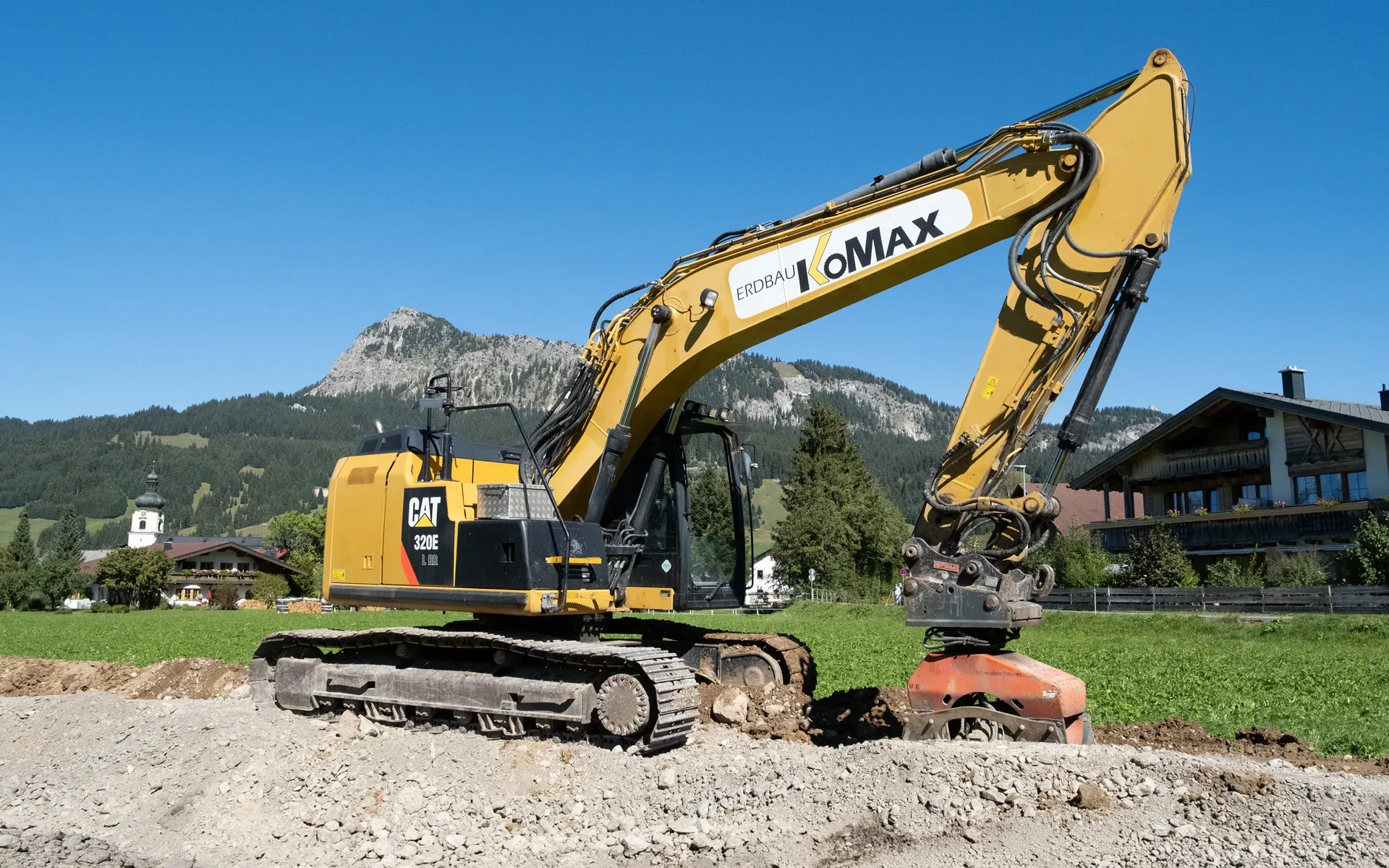 Gelber Cat-Kettenbagger mit KoMax-Schriftzug und Anbauverdichter auf einer Schotterfläche, dahinter Wiese, Kirchturm und Berg