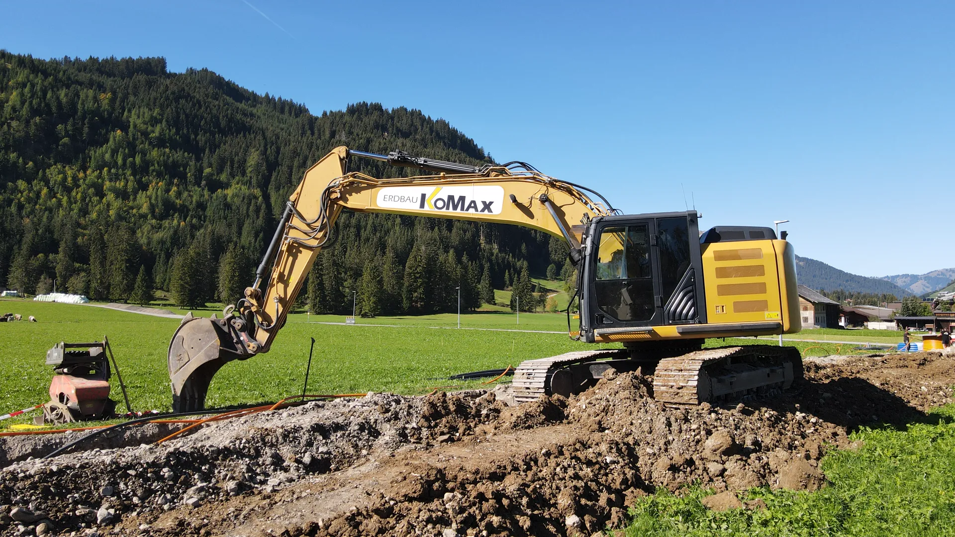 Gelber KoMax-Kettenbagger mit Tieflöffel auf frisch aufgeschüttetem Erdreich, dahinter Wiese und bewaldeter Berg