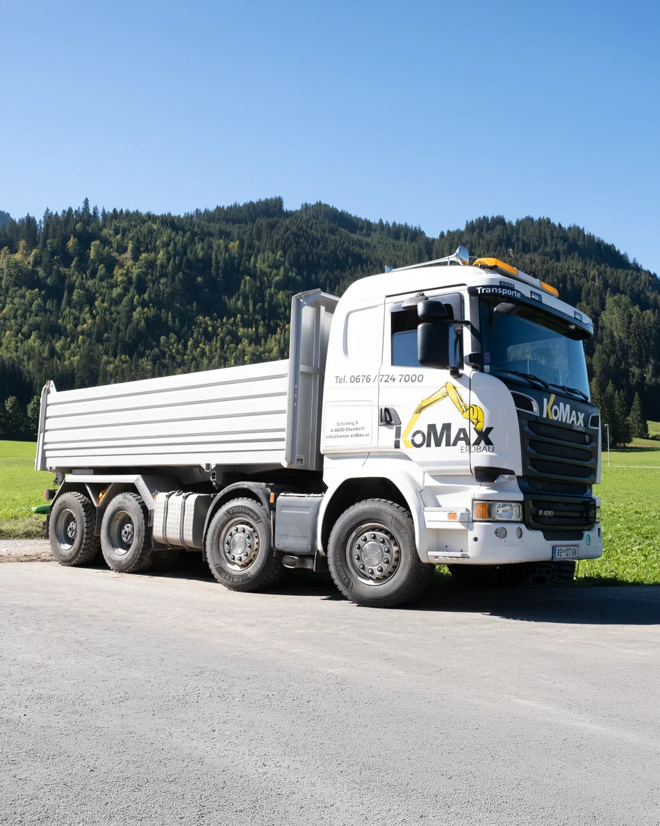 Weißer KoMax-Lkw mit Kipperaufbau auf einem Schotterweg vor Wiese und bewaldetem Berg