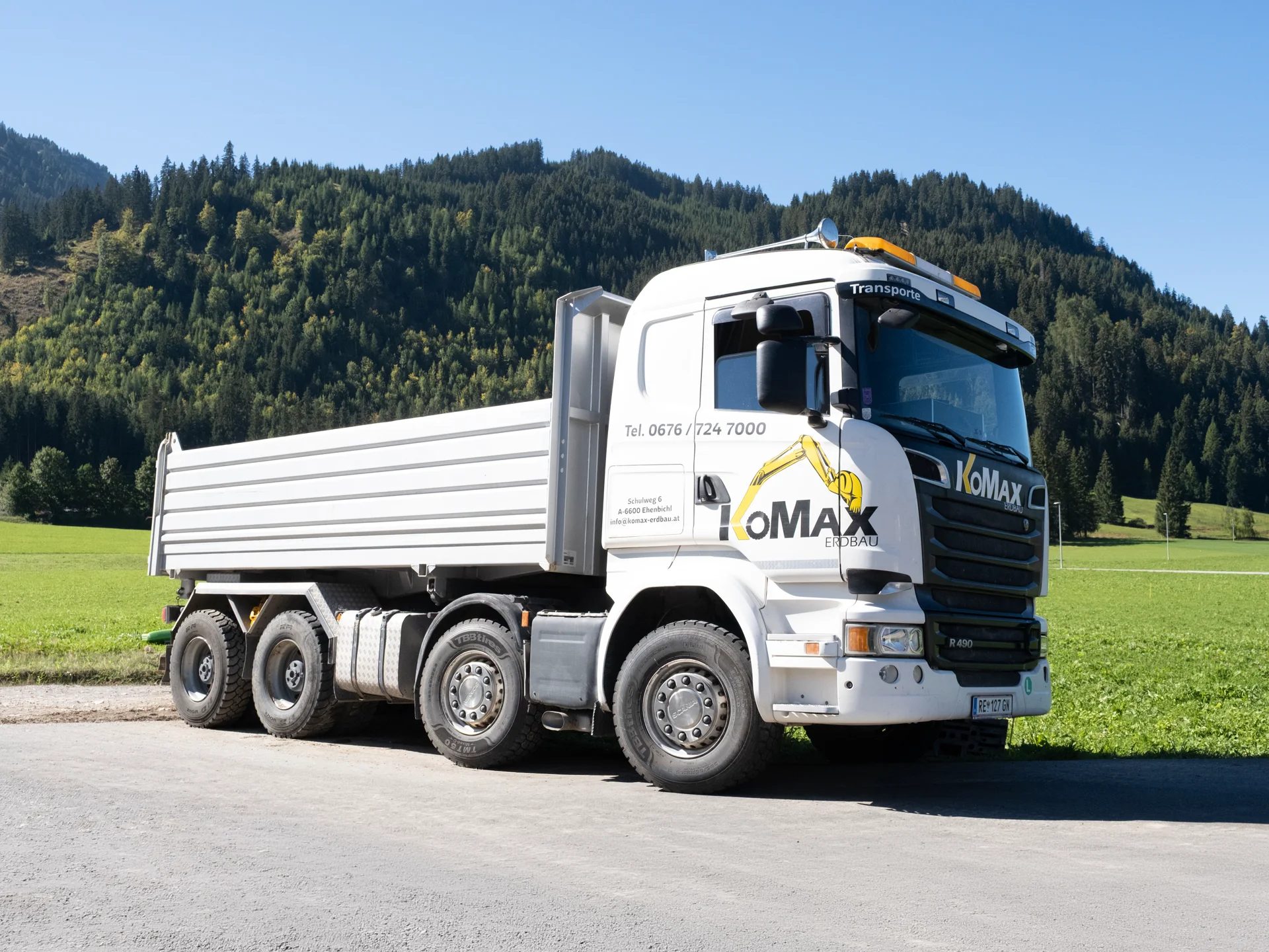 Weißer KoMax-Lkw mit Kipperaufbau von schräg vorne auf einem Schotterweg, dahinter Wiese und bewaldeter Berg