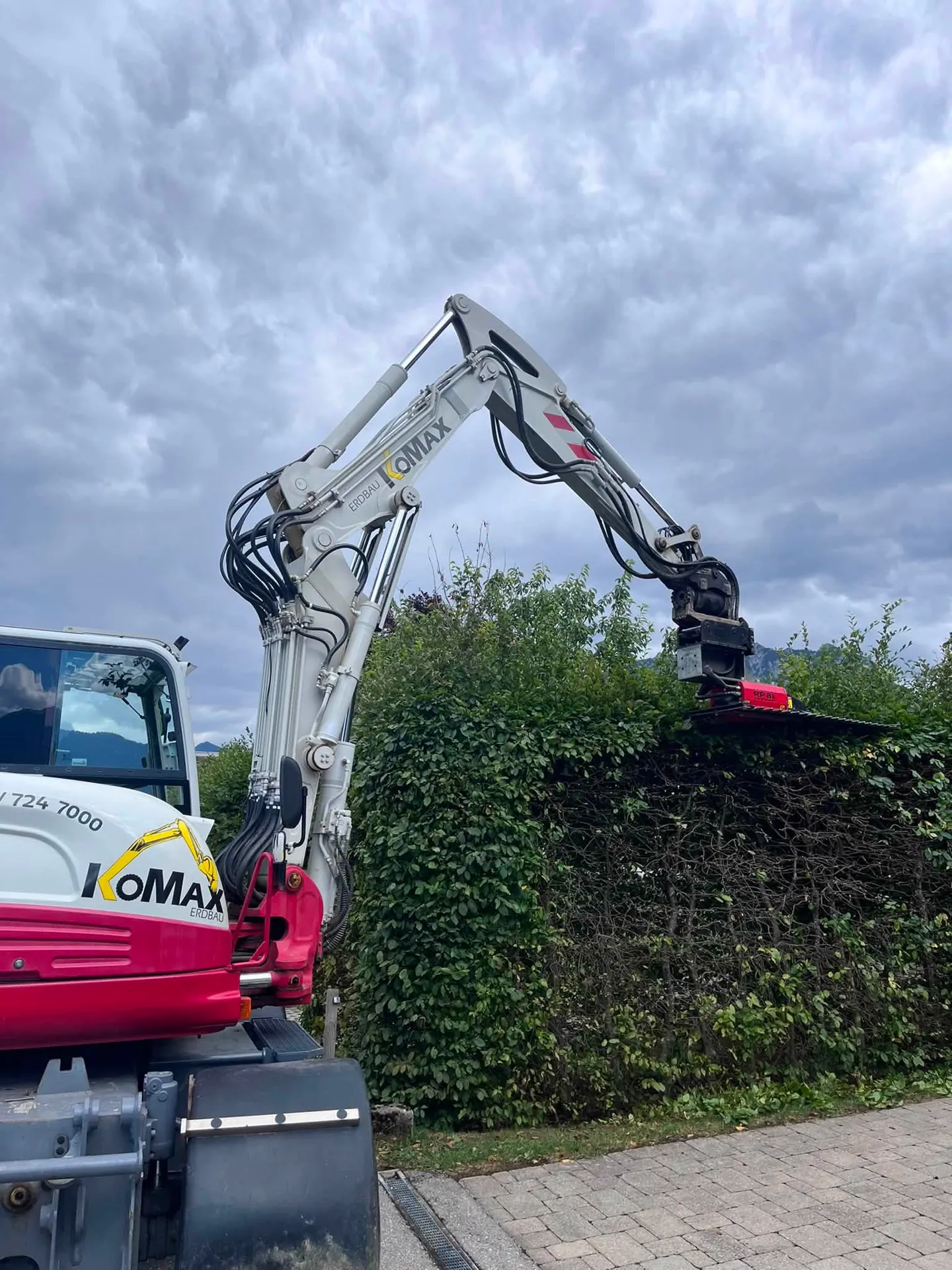Weiß-roter KoMax-Bagger mit Heckenschere schneidet eine hohe Hecke