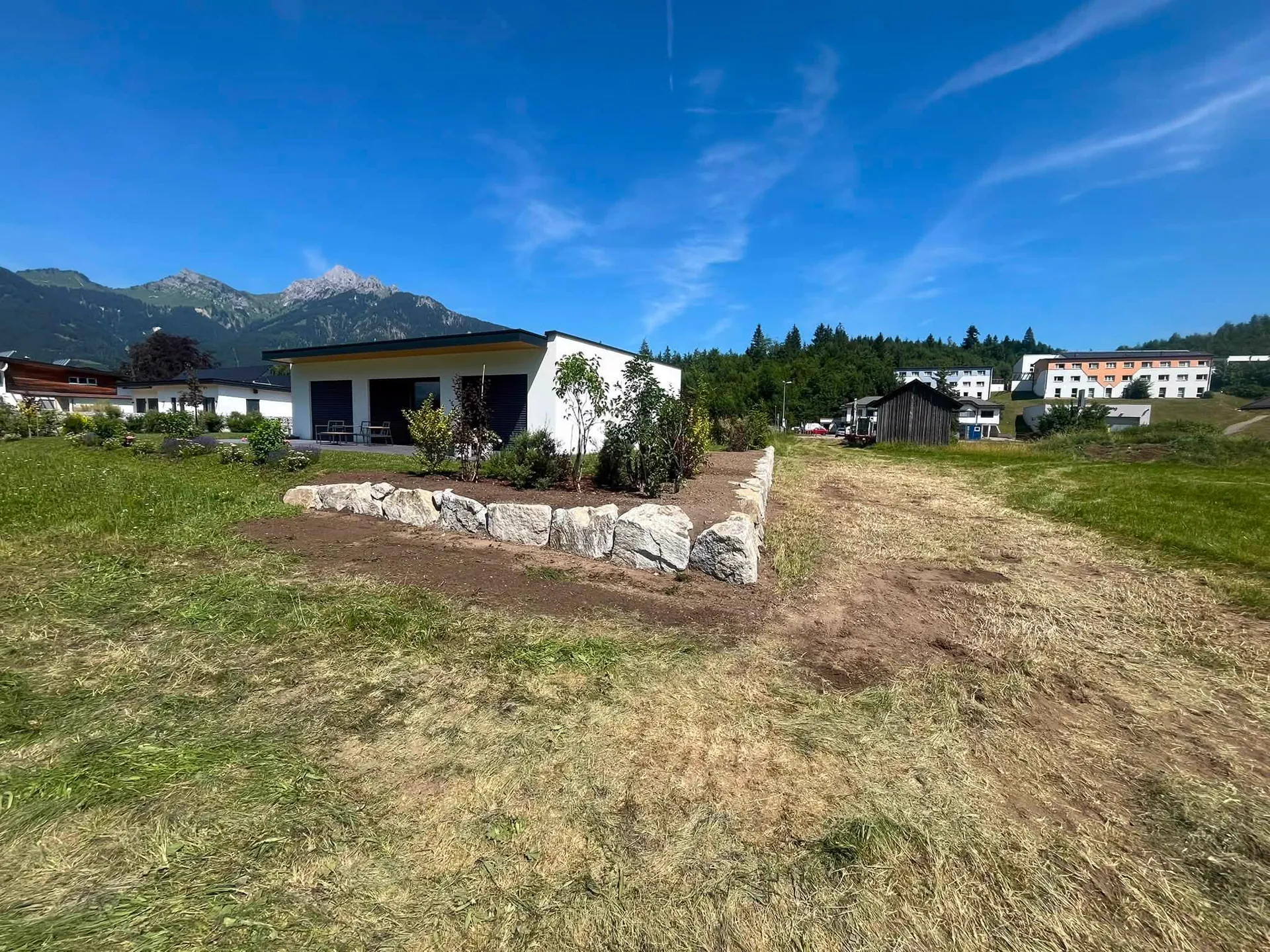 Neu angelegter Garten mit Natursteinmauer und jungen Bäumen vor einem Bungalow, dahinter die Berge
